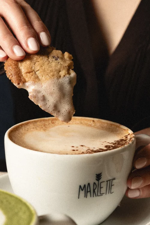 Main plongeant un biscuit bio fait-maison dans un café crème Marlette servi dans une tasse blanche