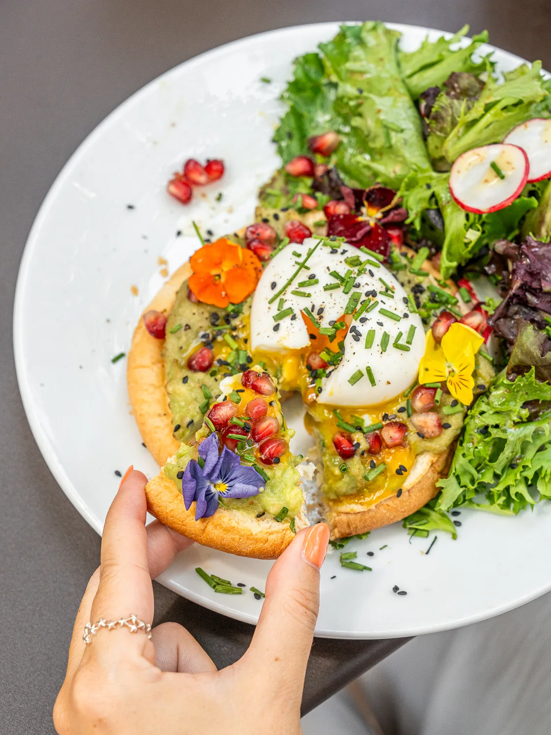 Main tenant une tranche de pain grillé avec avocat, œuf poché et fleurs comestibles, salade verte fraîche à côté, chez Marlette Martyrs à Pigalle