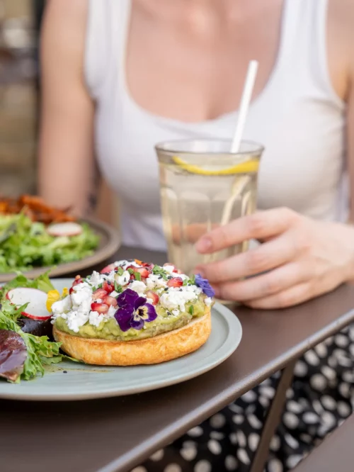 Cliente attablée tenant un verre d'eau citron devant son avocado toast garni de fleur violette et légumes frais au coffee shop Marlette des Abbesses, Montmartre
