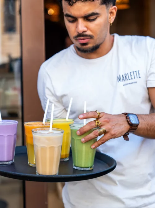 Barista en t-shirt blanc Marlette présentant un plateau de jus frais colorés aux Abbesses, Montmartre.