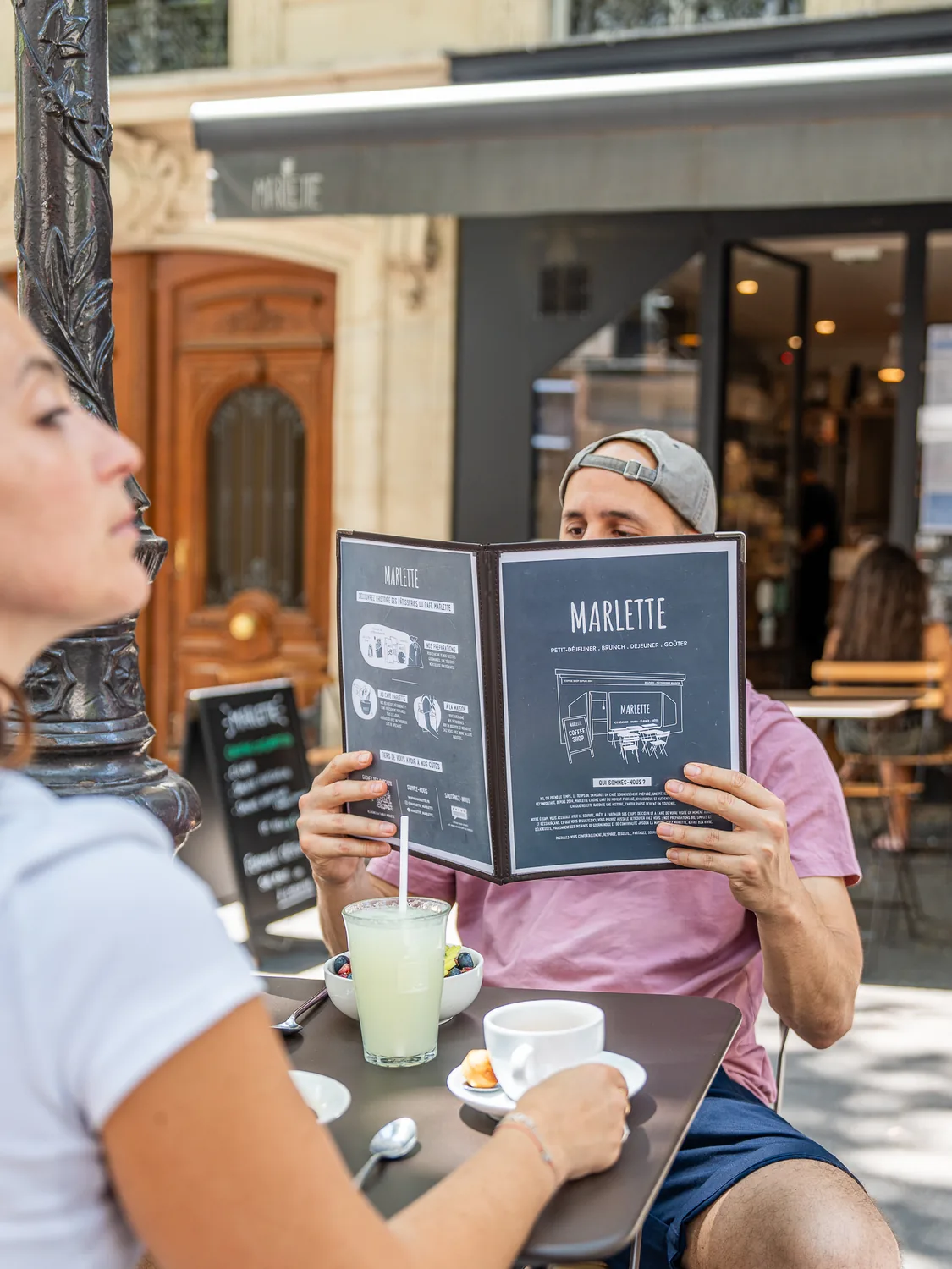 Deux clients assis à la terrasse du coffee shop Marlette rue des Martyrs, l'un consulte le menu tandis que l'autre déguste une boisson fraîche, avec la devanture du café en arrière-plan à Pigalle.