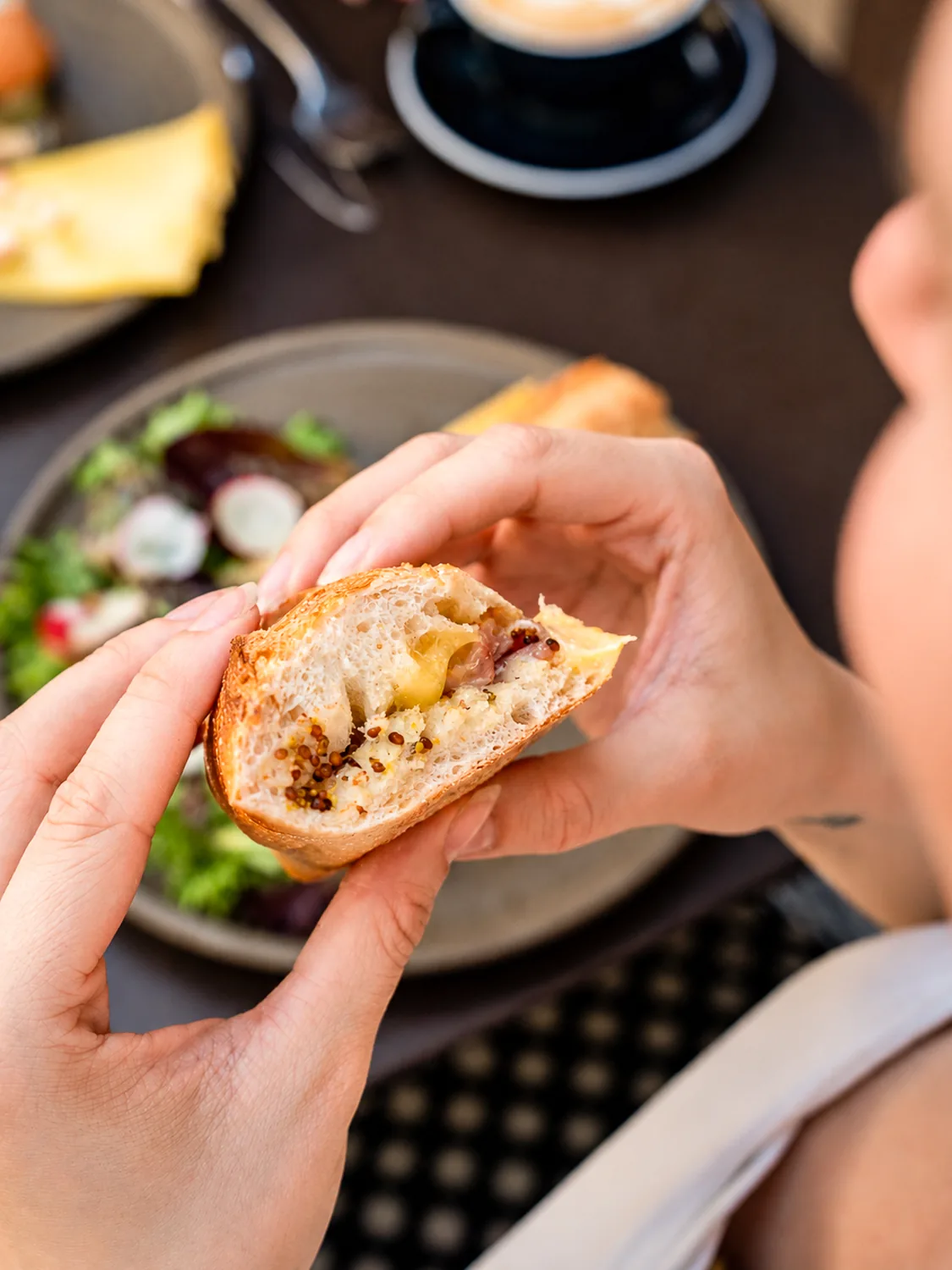 Client tenant une brioche briochée et garnie dans les mains, au coffee shop Marlette des Abbesses à Montmartre, avec salade et café en arrière-plan