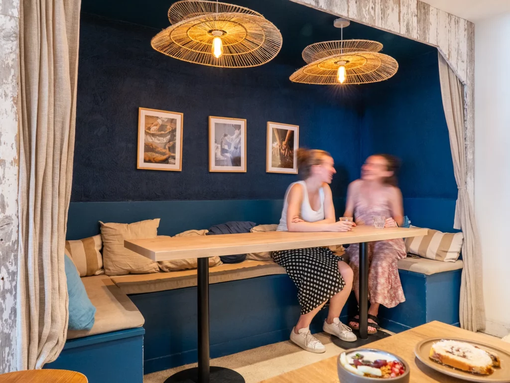 Deux clientes assises à une banquette bleue dans le coffee shop Marlette à Montmartre, sous des luminaires en rotin, avec assiettes de brunch visible au premier plan