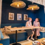 Deux clientes assises à une banquette bleue dans le coffee shop Marlette à Montmartre, sous des luminaires en rotin, avec assiettes de brunch visible au premier plan