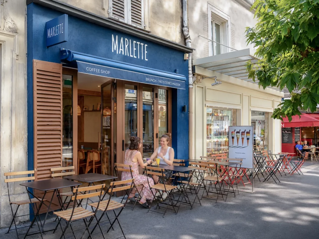 Façade bleue du coffee shop Marlette aux Abbesses avec terrasse aménagée de chaises et tables en bois, deux clientes assises en terrasse, Montmartre