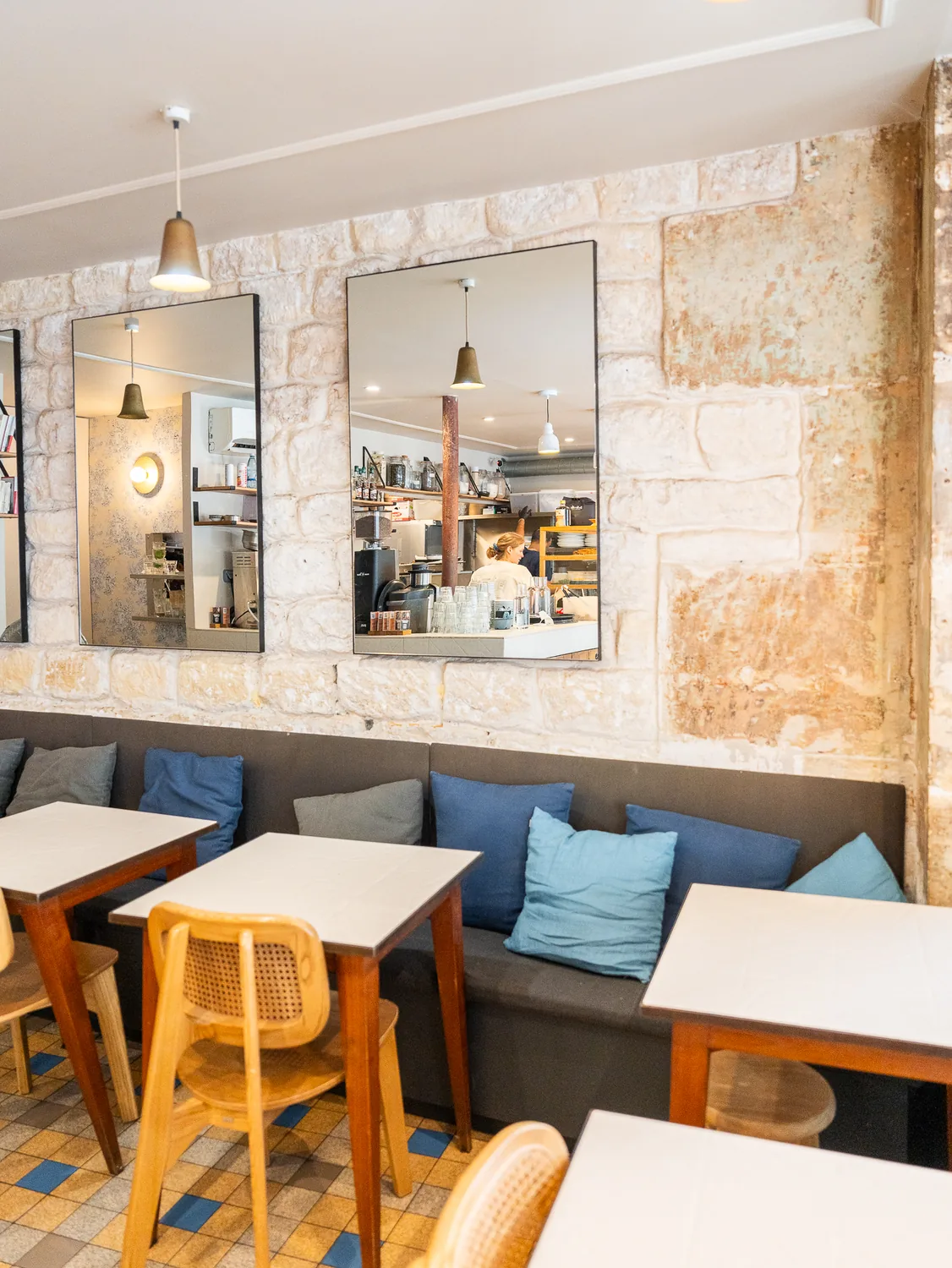 Intérieur du coffee shop Marlette à Pigalle avec banquette grise, coussins bleus, tables blanches, chaises jaunes en rotin et murs en pierre brute