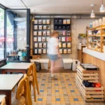 Intérieur spacieux du coffee shop Marlette à Pigalle avec comptoir en bois, étagères de produits bio, tables et chaises en bois naturel, client en mouvement