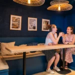 Deux clientes assises sur une banquette bleue dans le coffee shop Marlette à Montmartre, discutant autour d'une table en bois clair