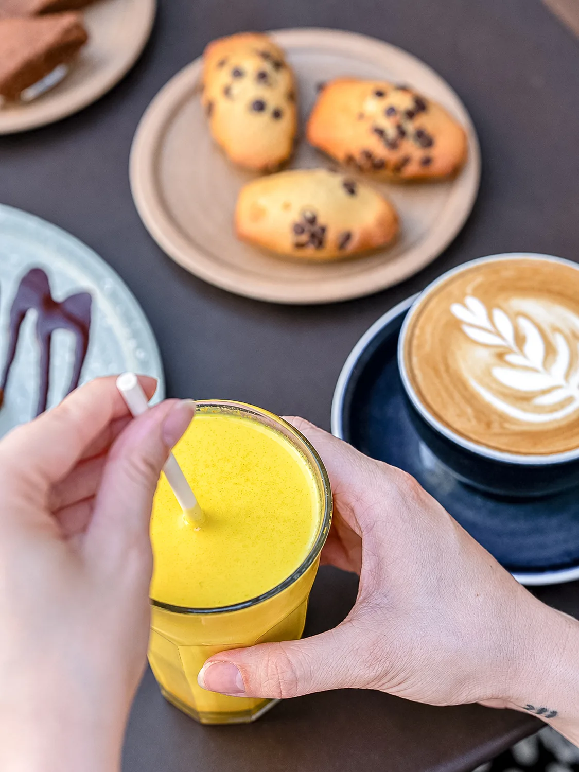 Main tenant un verre de jus frais jaune, assiette de cookies aux pépites de chocolat et cappuccino avec latte art chez Marlette à Montmartre