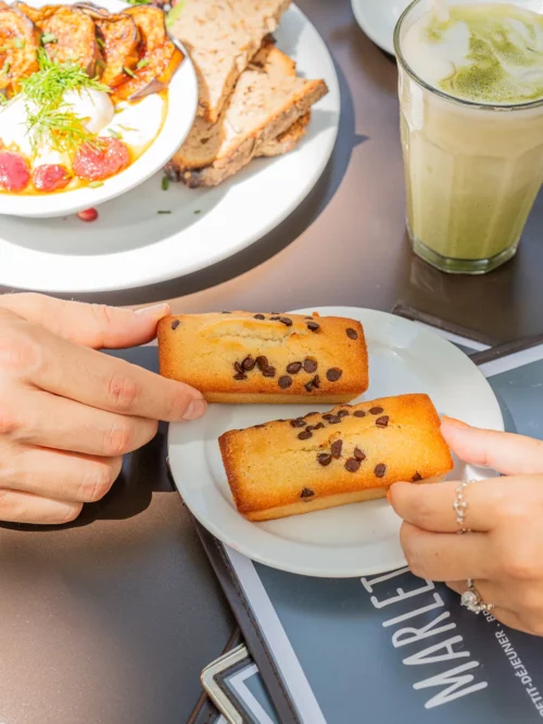Deux madeleines maison aux pépites de chocolat dans les mains d'une cliente au coffee shop Marlette des Martyrs à Pigalle, avec brunch et boisson en arrière-plan